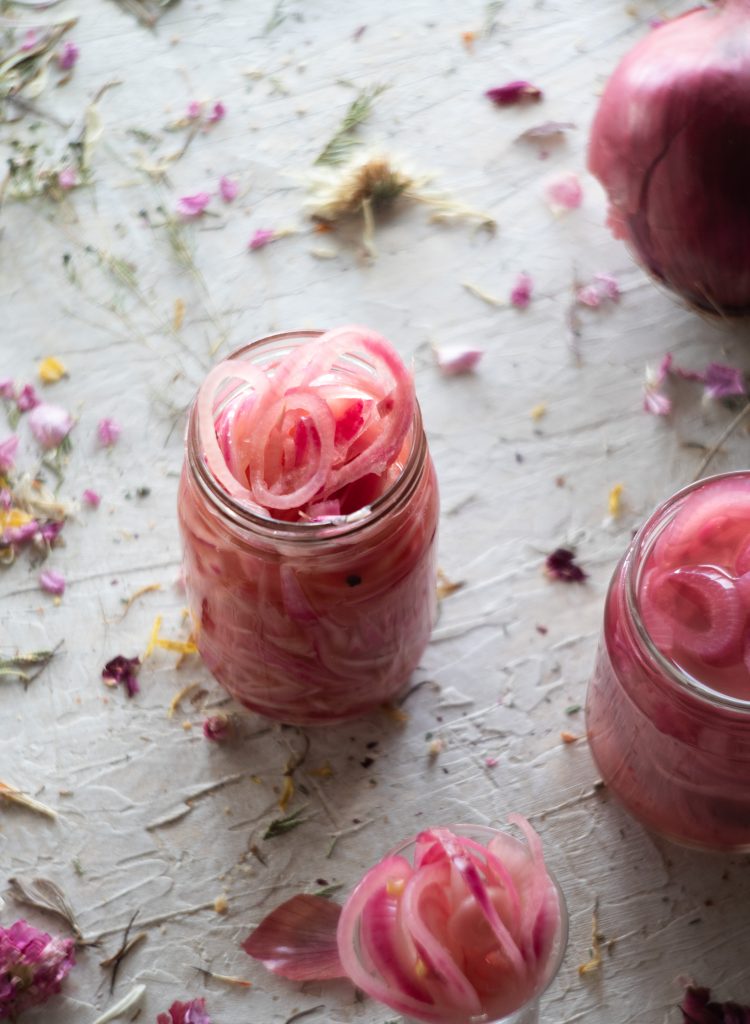 A jar of pickled red onion on a white rustic table with flower petals, with a red onion in the background and another jar of red onions in the forground. 
