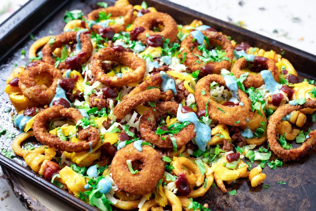 The vegan cheeseburger, onion ring, curly fry nachos on a metal sheet.