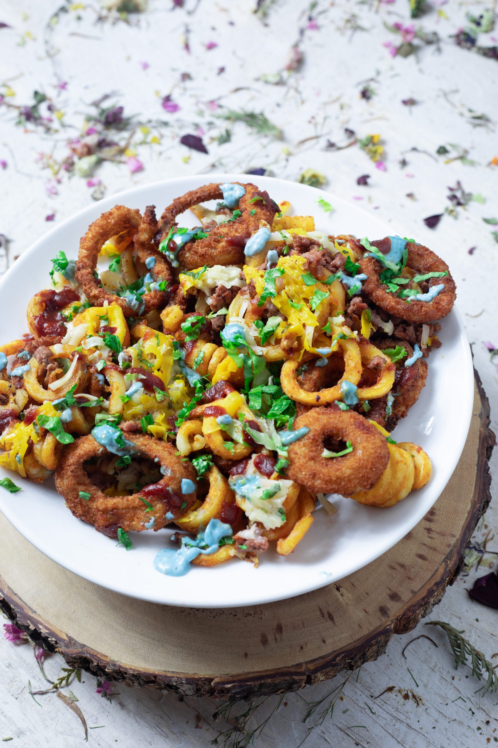 A plate of curly fries, then a layer of onion rings over the fries, then a layer of beefy crumbles, then a layer of melted plant cheddar cheese, then a layer of shredded lettuce, then a drizzle of katsup, then another drizzle of blue burger sauce