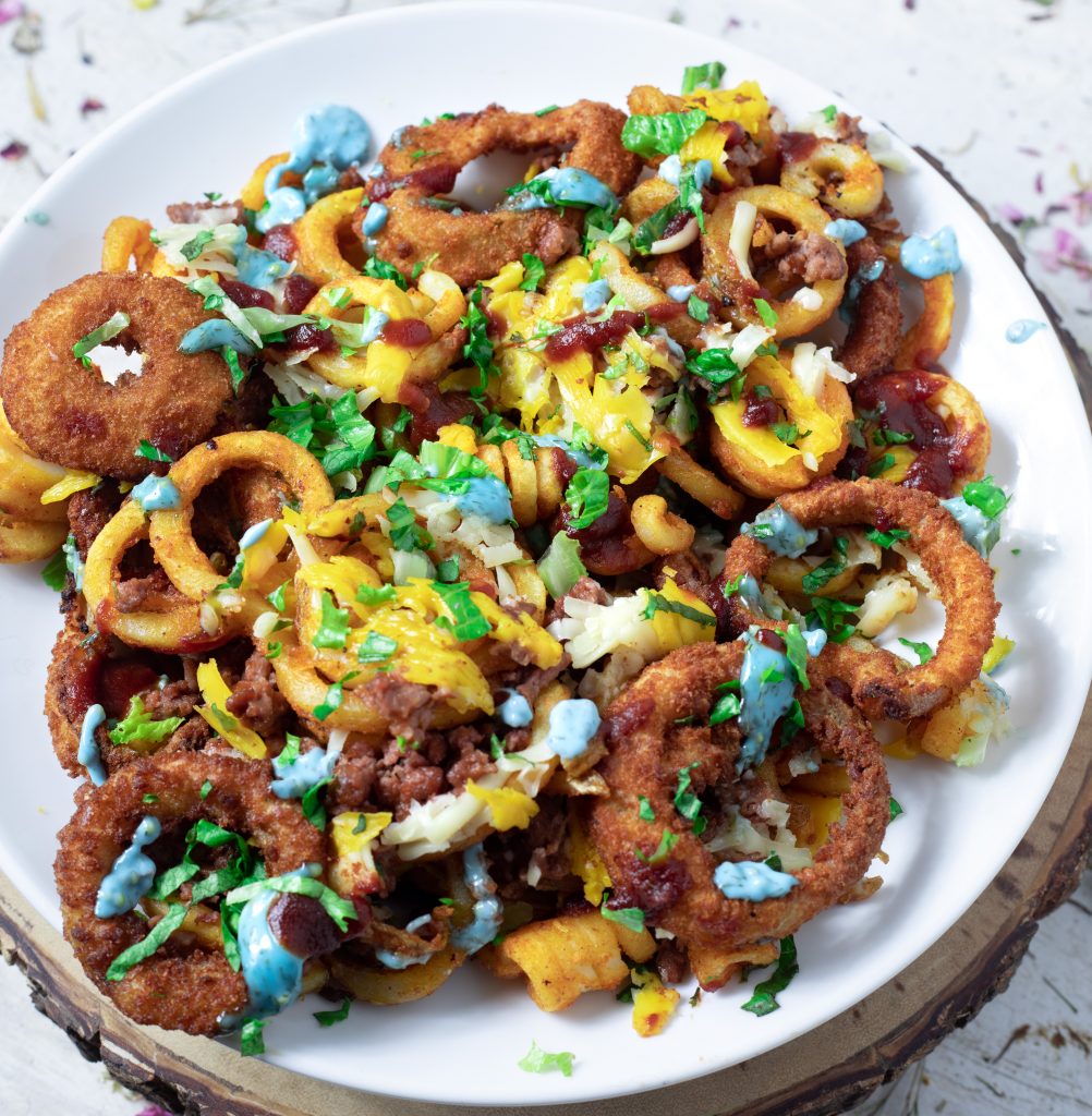 On a white plate sits a pile of curly fries, onion rings, vegan cheddar cheese, blue burger sauce, with bits of beyond burger poking through. 