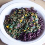 A geometic styled white bowl has a bed of black and brown rice, resulting in a purple hue. Layered over the purple rice rest hot spinach and purple potato curry with yellow flower petals sprinked over the top.