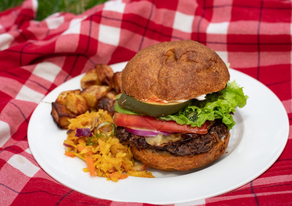 A veggie burger with pickles, lettuce, tomato, and grilled onions on a whole wheat bun is sitting next to a pile of orange kohlrabi golden beet slaw with roasted potatoes behind. 