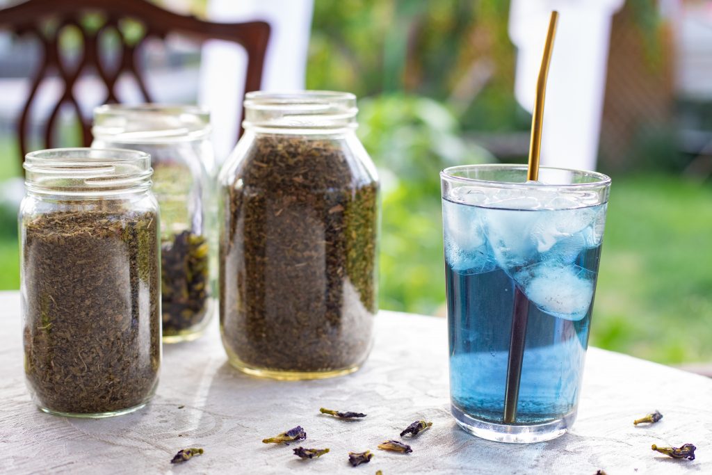 A glass of mermaid mint iced tea on a table with jars of herbs behind. 