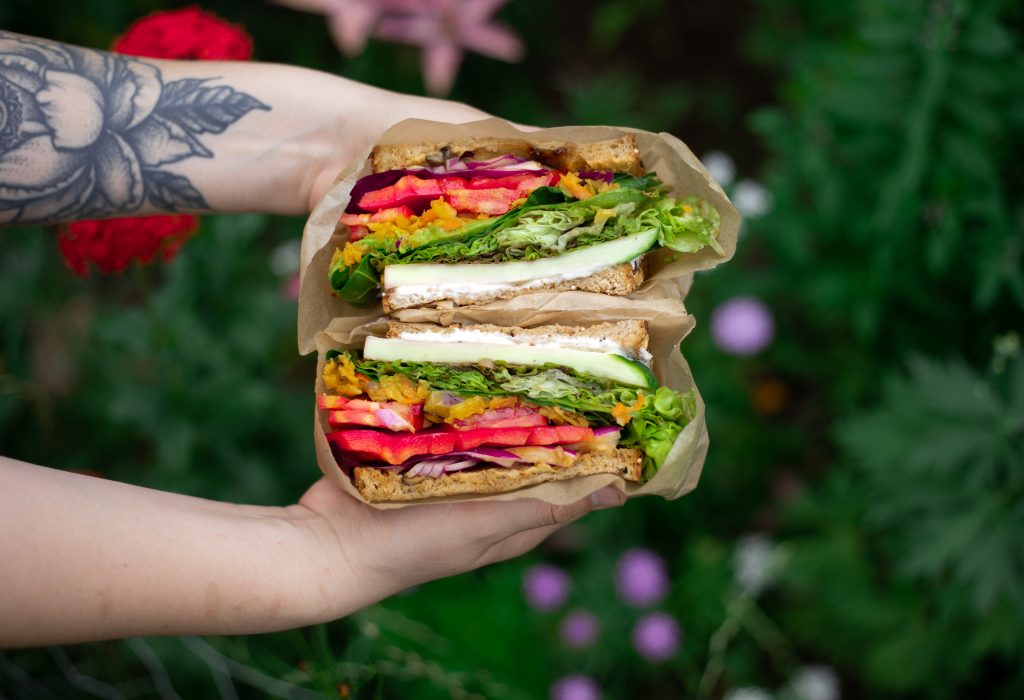 A sandwich wrapped in parchment paper being held in front of flowers. The sandwich is layered with vegetables in colors ranging from purple, red, orange, green and white