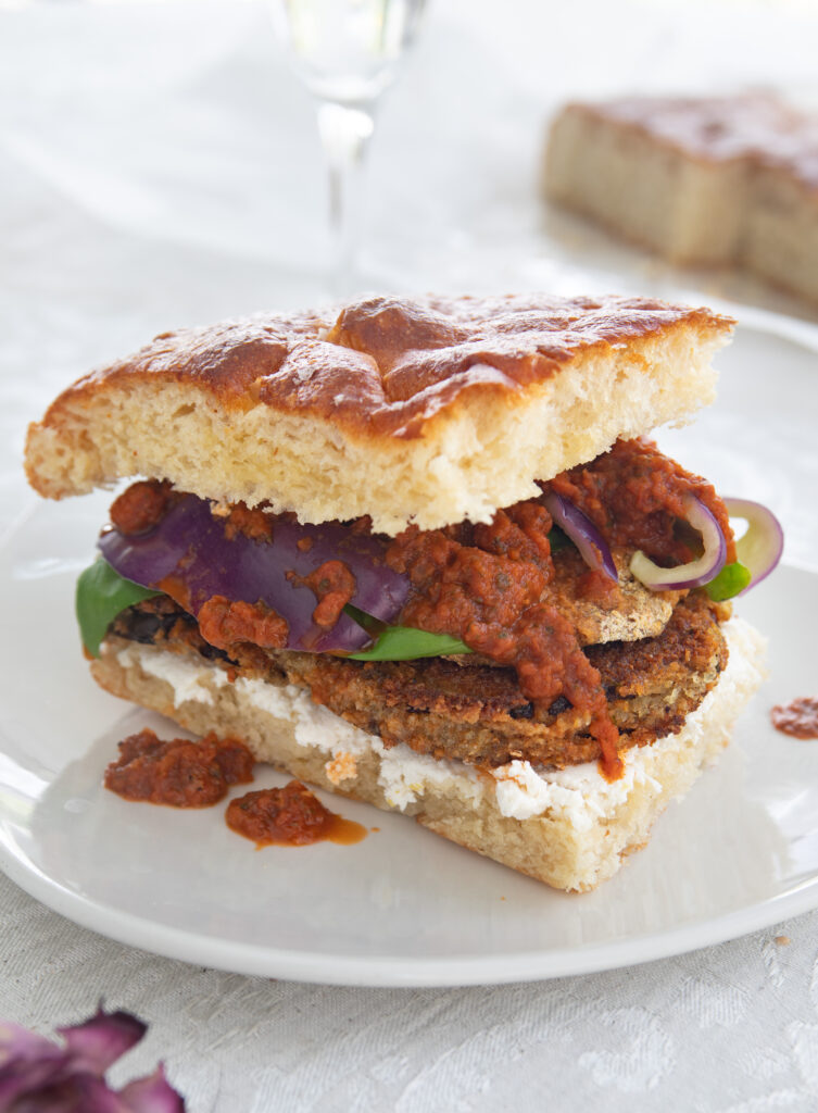 A sandwich made of focaccia bread with red sauce, breaded eggplant, fresh basil, purple bell pepper, and almond ricotta cheese. 