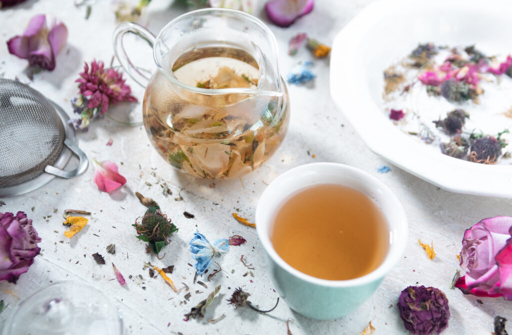 A glass tea pot filled with floral tea with a poured cup of tea next to it