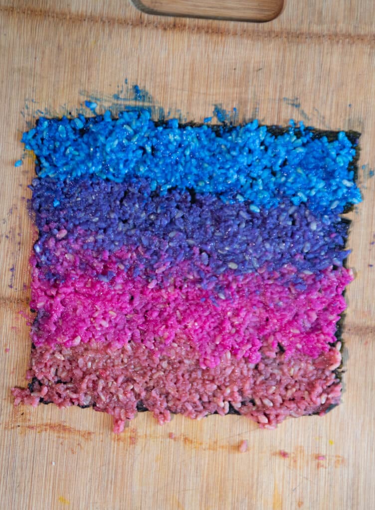 Colorful sushi rice on a nori sheet, the colors gradate from blue to pink. 