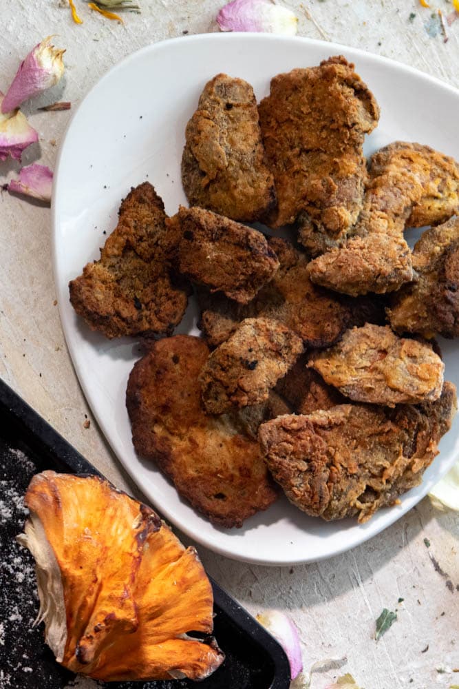 A plate of breaded and fried chicken of the woods next to a black sheet with a fresh chicken of the woods mushroom. 