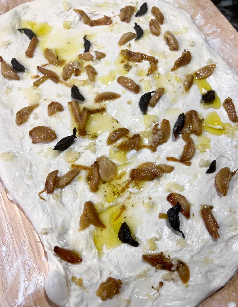Dough Stretched out into a rectangle covered in cloves of garlic confit, black garlic, with grated garlic and small dollops of garlic oil