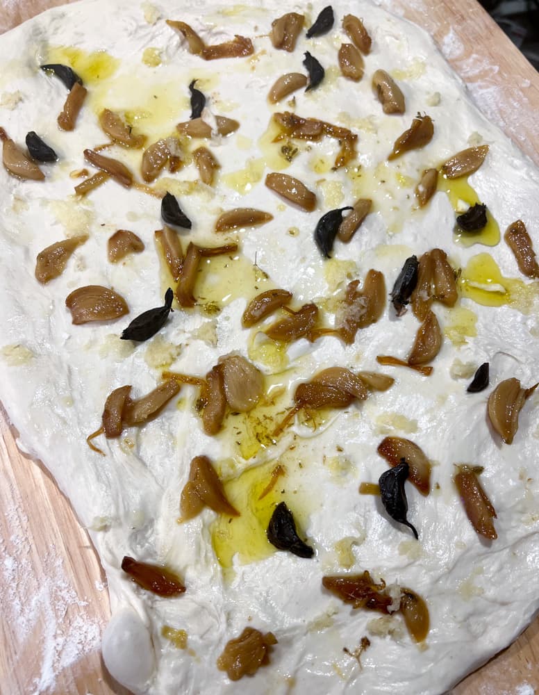 Dough Stretched out into a rectangle covered in cloves of garlic confit, black garlic, with grated garlic and small dollops of garlic oil 