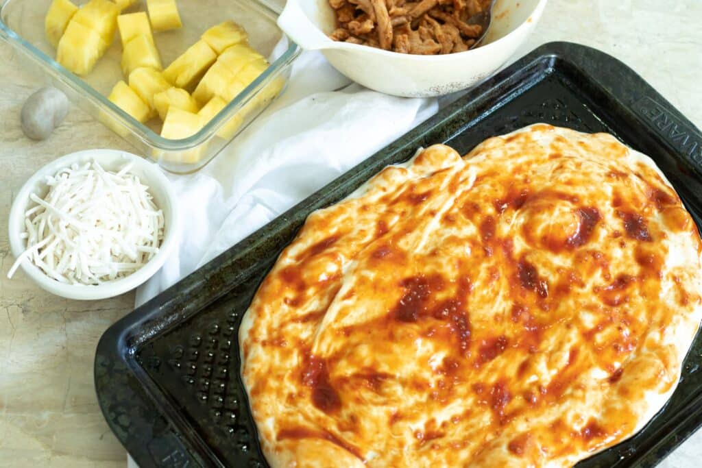 Pizza dough stretched out with bbq sauce next to cheese, pineapple, and soy curls 