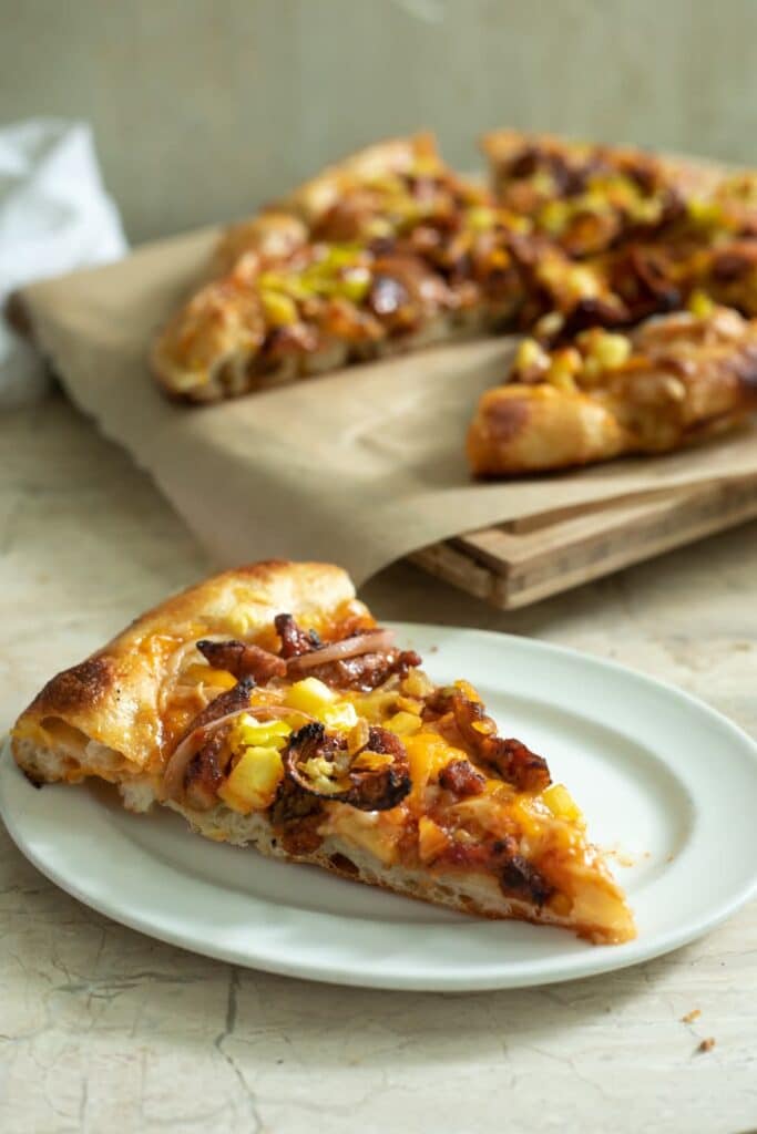 A slice of vegan bbq pizza on a white plate in front of the cut pizza