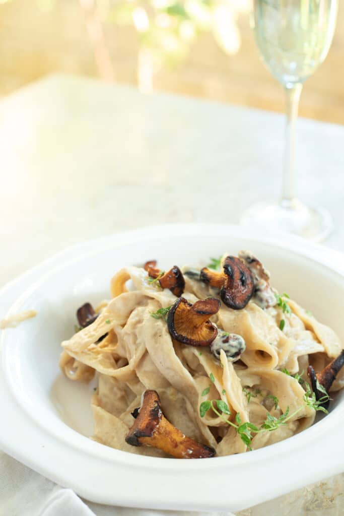 a plate of creamy pasta with chanterelle mushrooms on a white table with a champagne glass