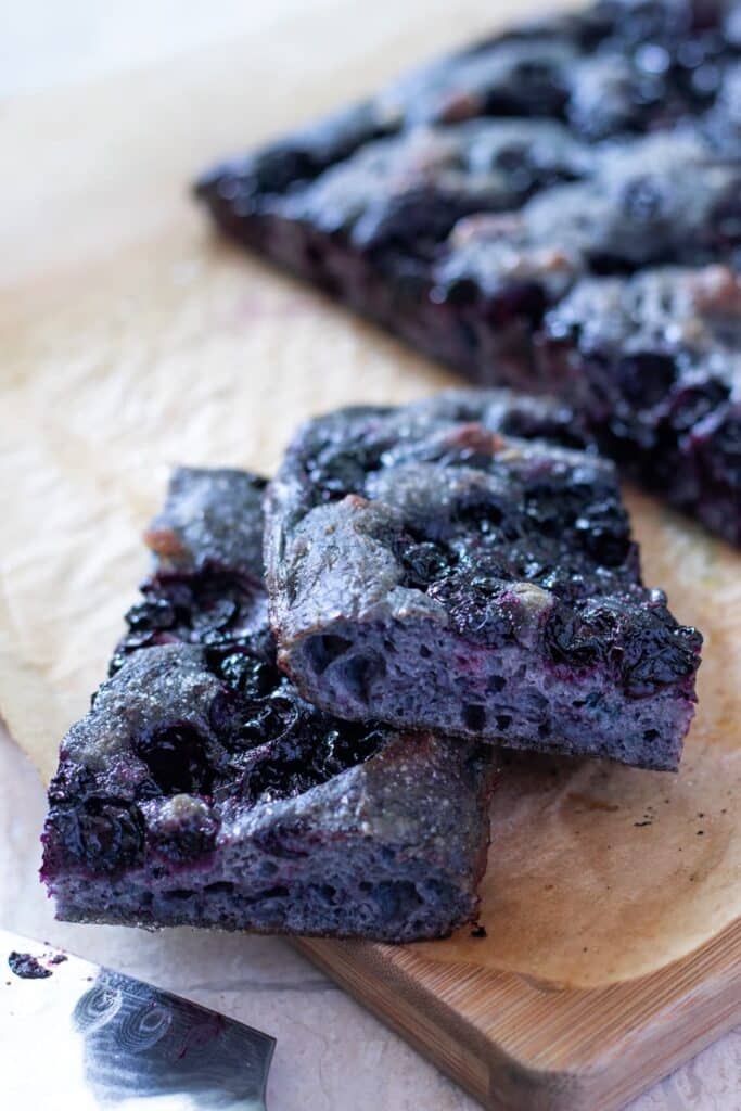 Two slices of blue sourdough focaccia bread that has blueberries baked in on a cutting board. 