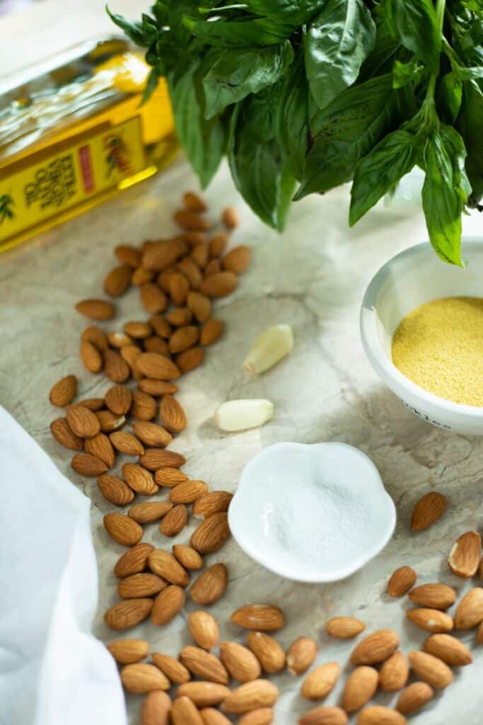 Almonds, Salt, Garlic, a small bowl of nutritional yeast flakes, fresh basil, and a bottle of olive oil sitting on a marble countertop