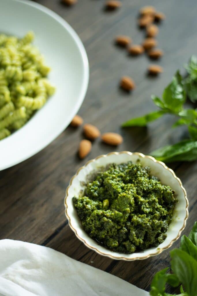 a small bowl of pesto on a wooden table next to a plate of pesto pasta