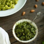 a small bowl of pesto on a wooden table next to fresh basil, almonds, and a plate of pesto pasta