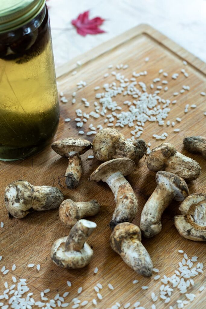 A wooden cutting board with uncooked short grain rice, dirt covered matsutake mushrooms, next to a glass jar filled with dashi 