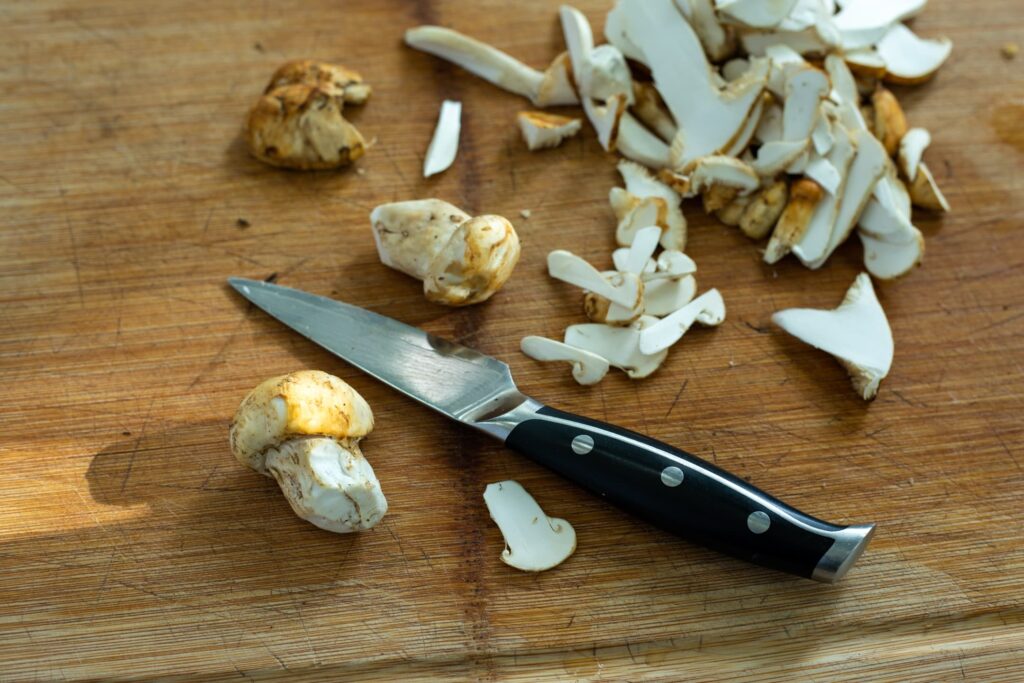 A wooden cutting board filled with cleanly sliced matsutake mushrooms with a knife and a couple of cleaned uncut mushrooms 