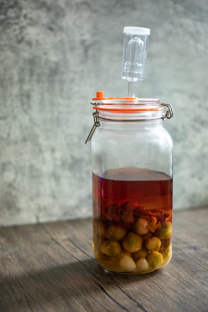 A glass fermentation jar filled with a pink brine and brussels sprouts.