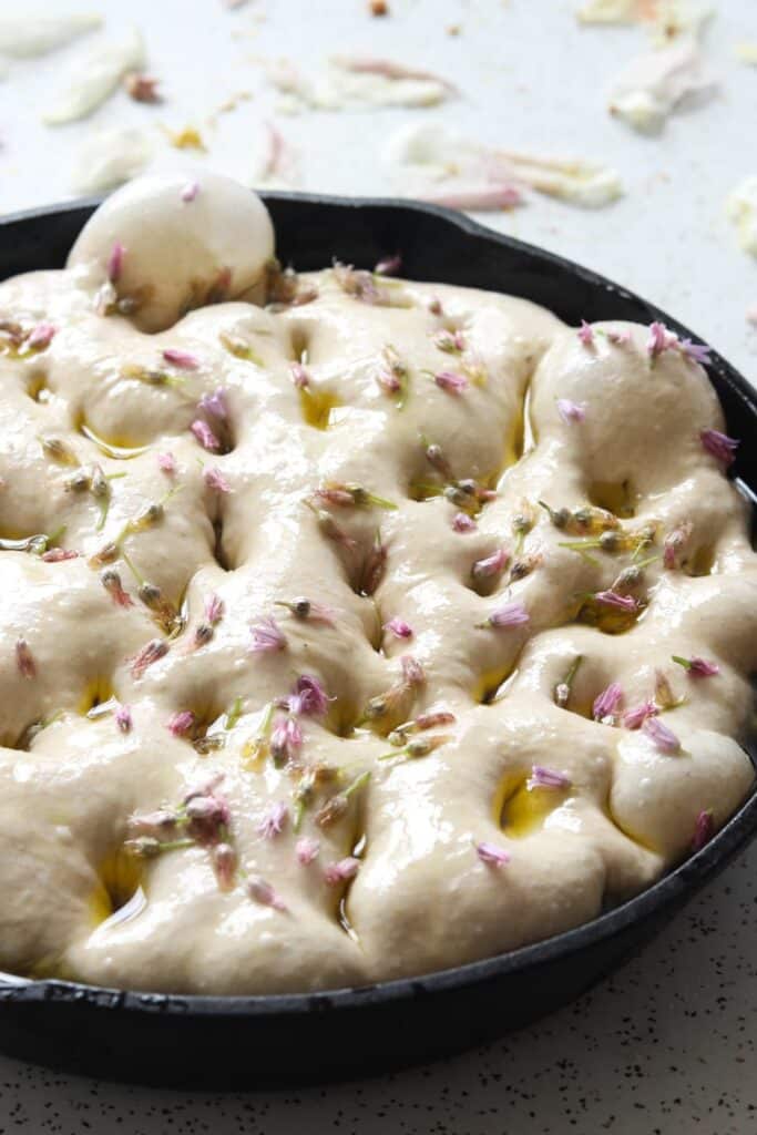 a raw focaccia topped with chive flowers in a cast iron skillet on a white table