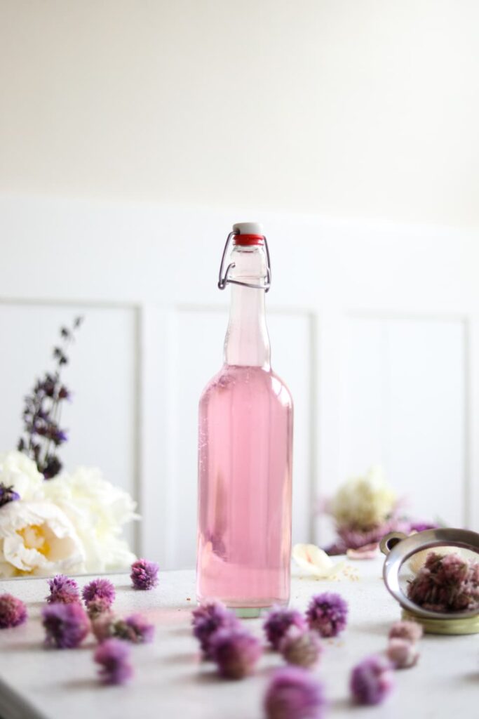 a glass jar filled with pink chive blossom vinegar on a table next to fresh chive blossoms