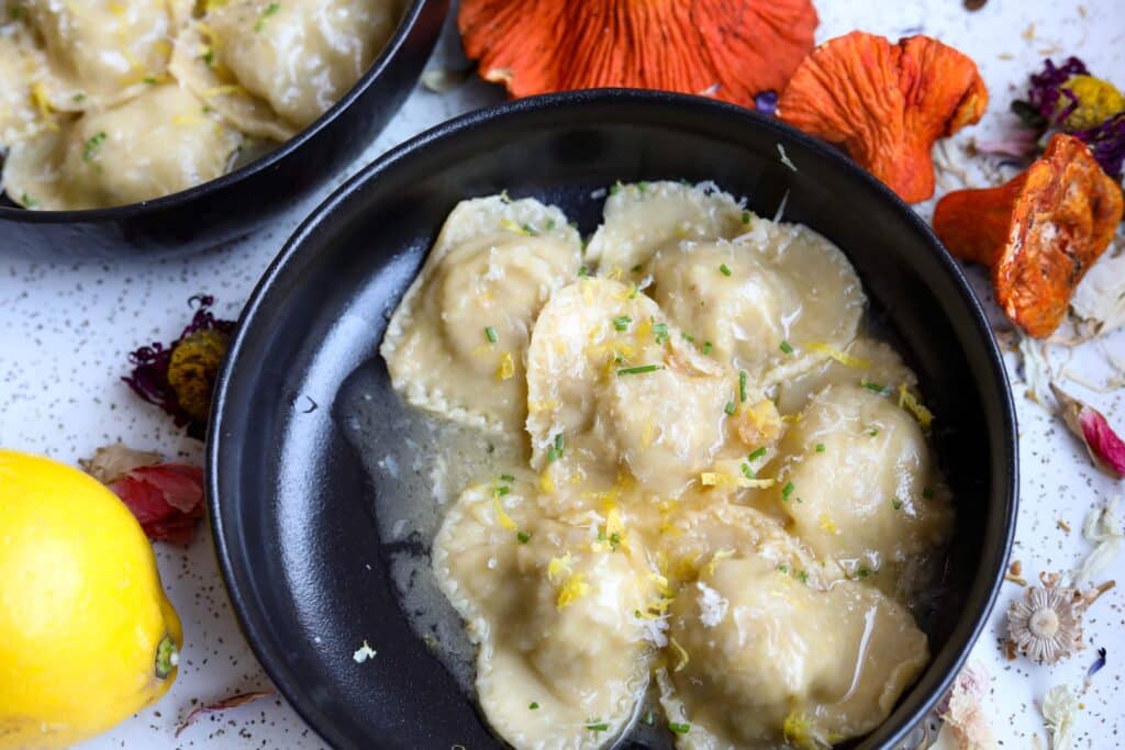 a black plate of ravioli in a sauce with lemon zest, chives, and vegan parmesan garnishing it